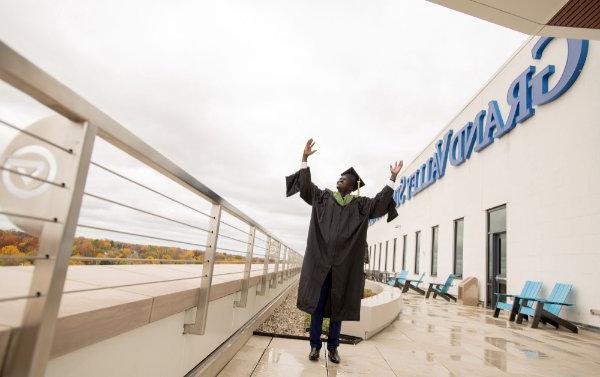 A person wearing a cap and gown has their arms in the air. The words Grand Valley State are visible in the background.