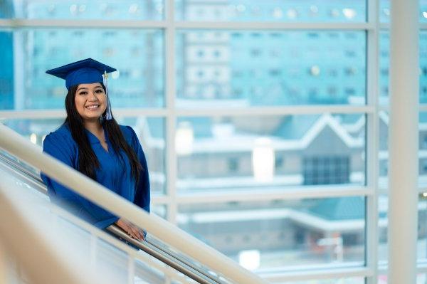 A person wearing a cap and gown smiles while standing on a staircase in front of windows with buildings in the background.
