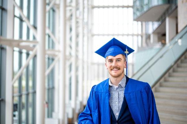 A student wearing a cap and gown smiles while standing in front of a stairway.