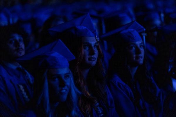  一段视频中的蓝光照射在戴着蓝色帽子、身穿蓝色长袍的毕业生脸上. 