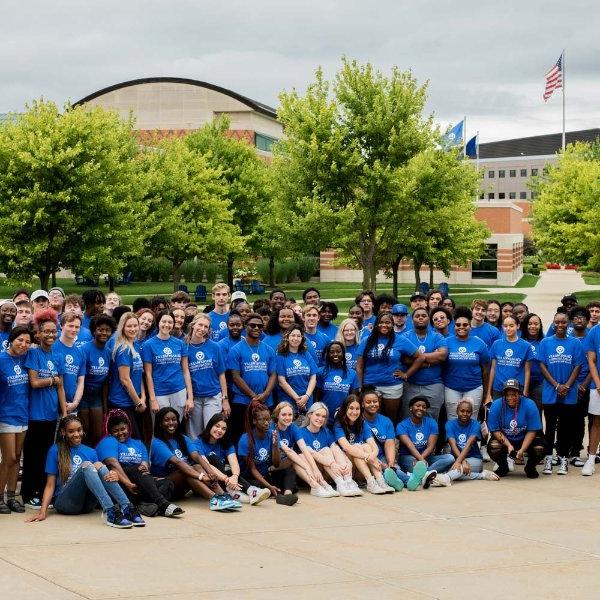 庞大的学生群体, faculty and staff all wearing blue GVSU Oliver Wilson Scholar t-shirts; pictured with Student Services Building behind them